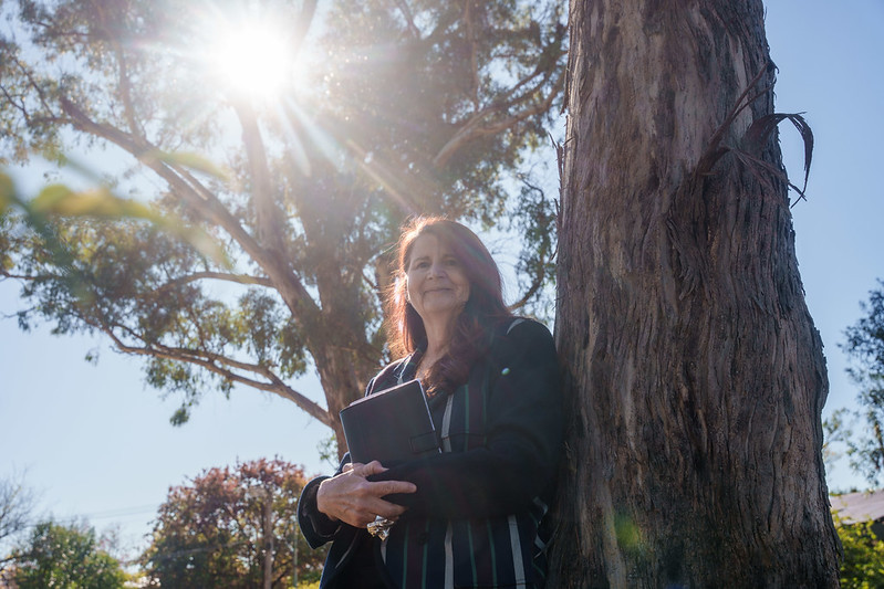 A poet with long red hair leans against a tree holding a book. The sun shines through a tall eucalyptus tree behind her.